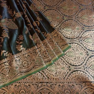 Banarasi Satin silk saree in Dark Bottle Green shade dual tone with peacock and creeper motifs in light gold zari weaves all over the body. Very unique meenakari work done on the motifs in the base fabric shade. It has been woven in the most famous jaal weaves i.e. All the motifs are connected throughout the saree. It has a self border with gold work floral and peacock motifs. The Pallu is richly woven with intricate Banarasi floral motifs. Meenakari work has been done In the border and pallu that compliments the whole look of the saree. It includes self embossed blouse piece with gold border in same design as the saree.