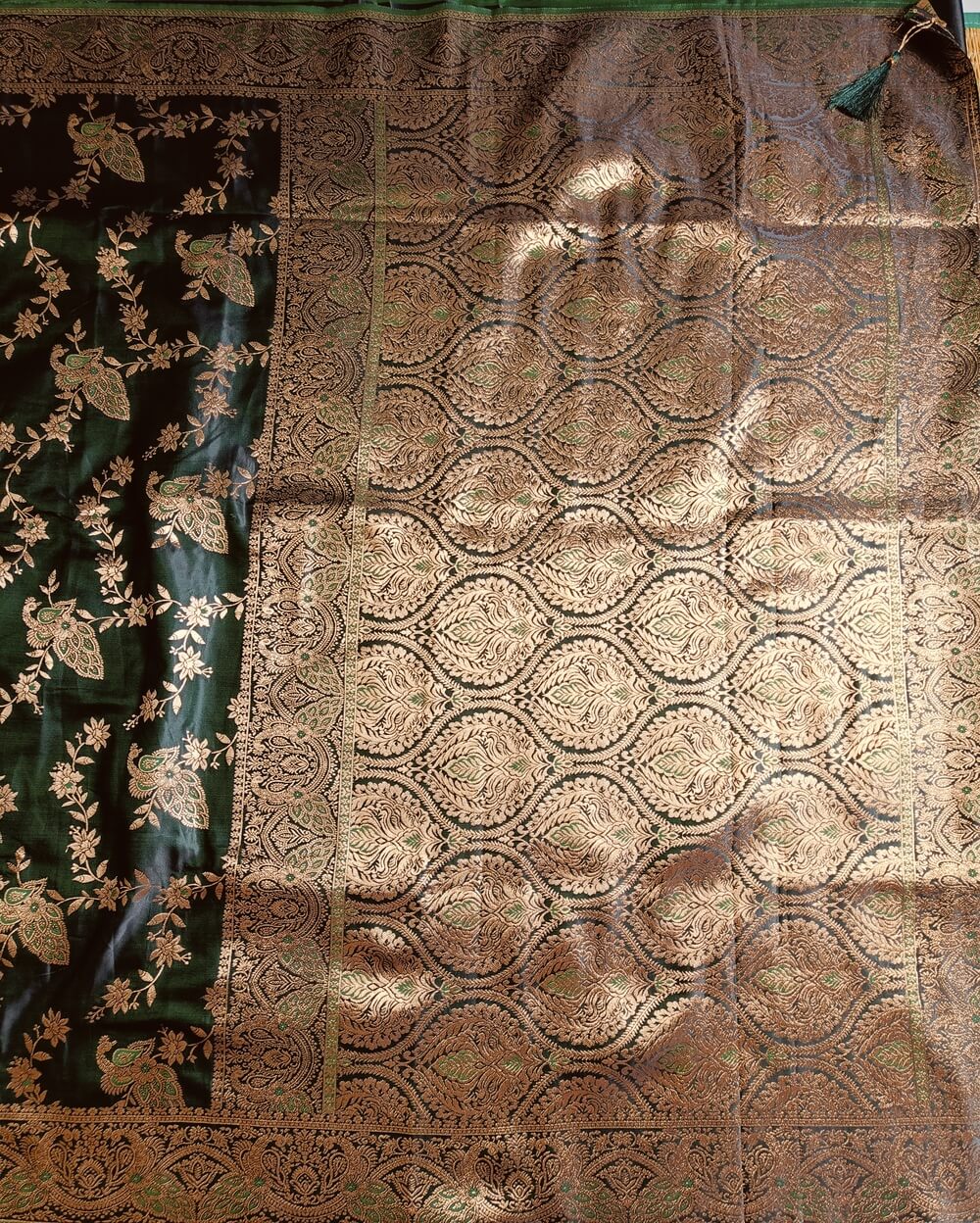 Banarasi Satin silk saree in Dark Bottle Green shade dual tone with peacock and creeper motifs in light gold zari weaves all over the body. Very unique meenakari work done on the motifs in the base fabric shade. It has been woven in the most famous jaal weaves i.e. All the motifs are connected throughout the saree. It has a self border with gold work floral and peacock motifs. The Pallu is richly woven with intricate Banarasi floral motifs. Meenakari work has been done In the border and pallu that compliments the whole look of the saree. It includes self embossed blouse piece with gold border in same design as the saree.